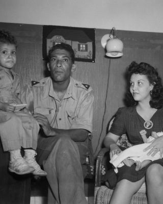 Een KNIL-militair zit met een vrouw en twee kinderen in een kamer.