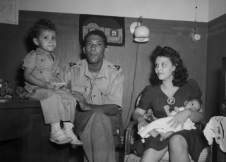 Een KNIL-militair zit met een vrouw en twee kinderen in een kamer.